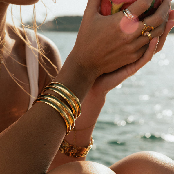 Person holding a dragon fruit by a body of water. Wearing gold plated cuff bracelets. 