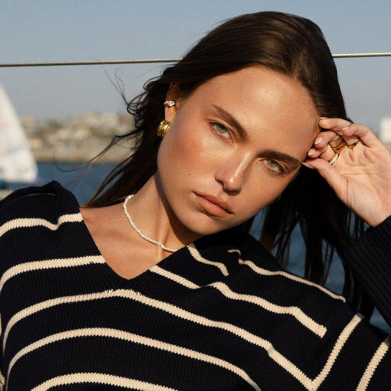 Woman wearing a black and white striped sweater with a scenic background wearing petite pearl necklace and vintage inspired gold earrings