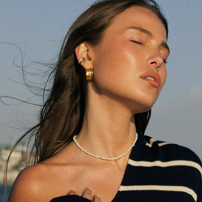Woman in a black and white striped sweater and white bikini sitting on a boat with water and sky in the background. wearing waterproof jewelry and pearl choker