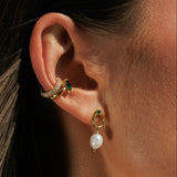 Close-up of an ear wearing gold earrings with green gemstones and pearl drops.