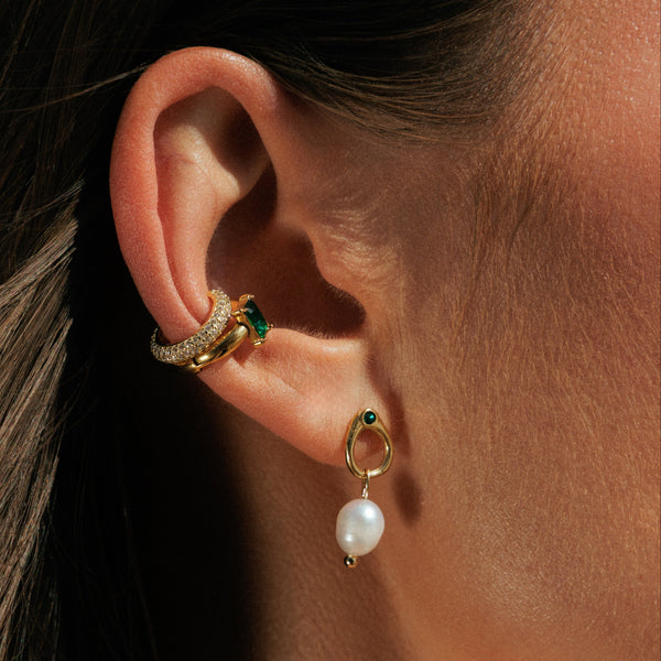 Close-up of an ear wearing gold earrings with green gemstones and pearl drops.